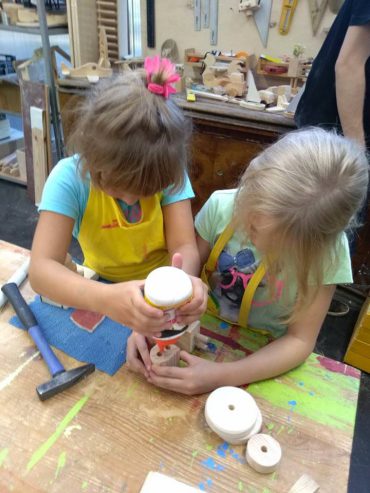 dwie dziewczynki kleją drewniane elementy podczas warsztatów